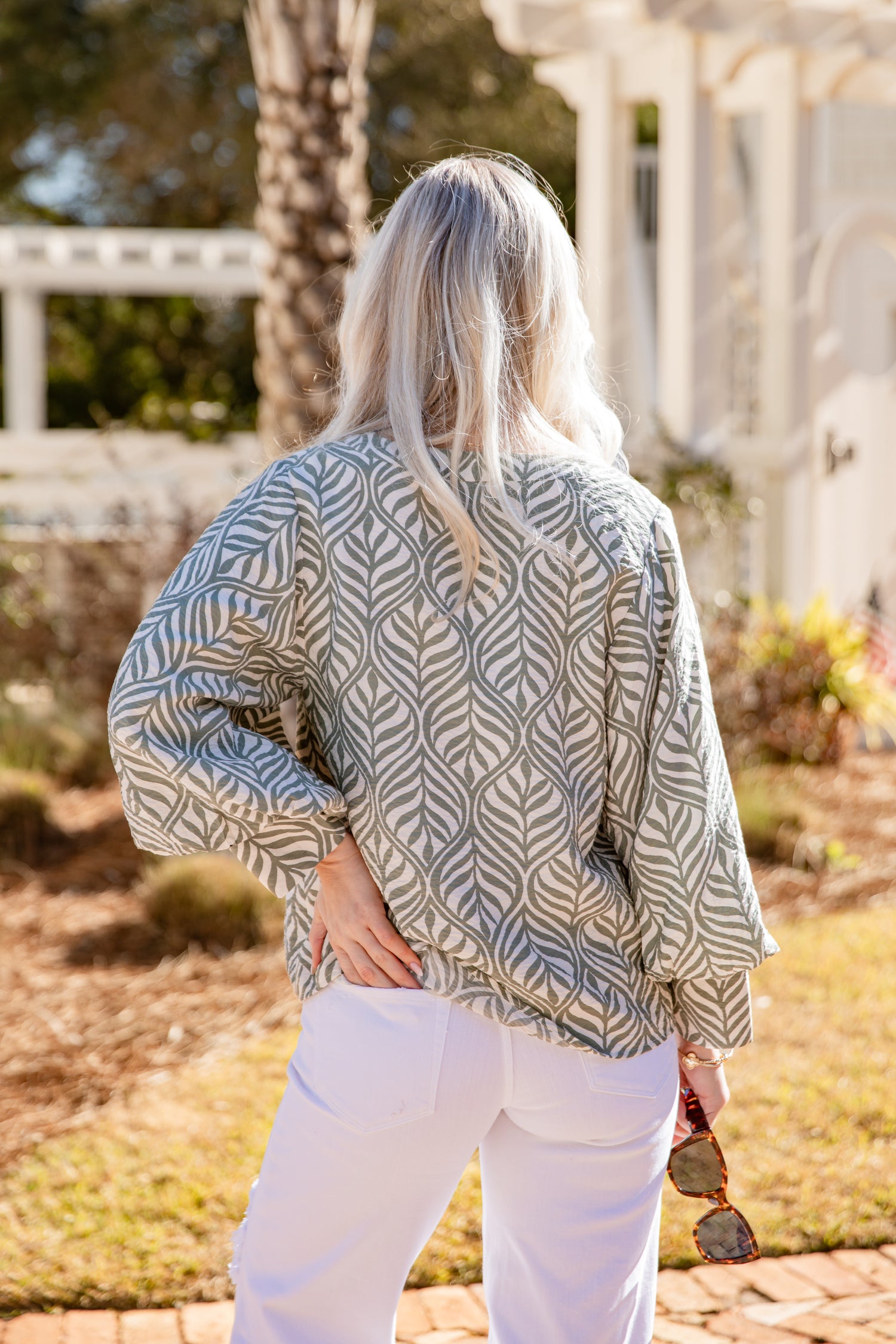 Woman wearing a patterned top and white pants standing outdoors with trees and a building in the background.