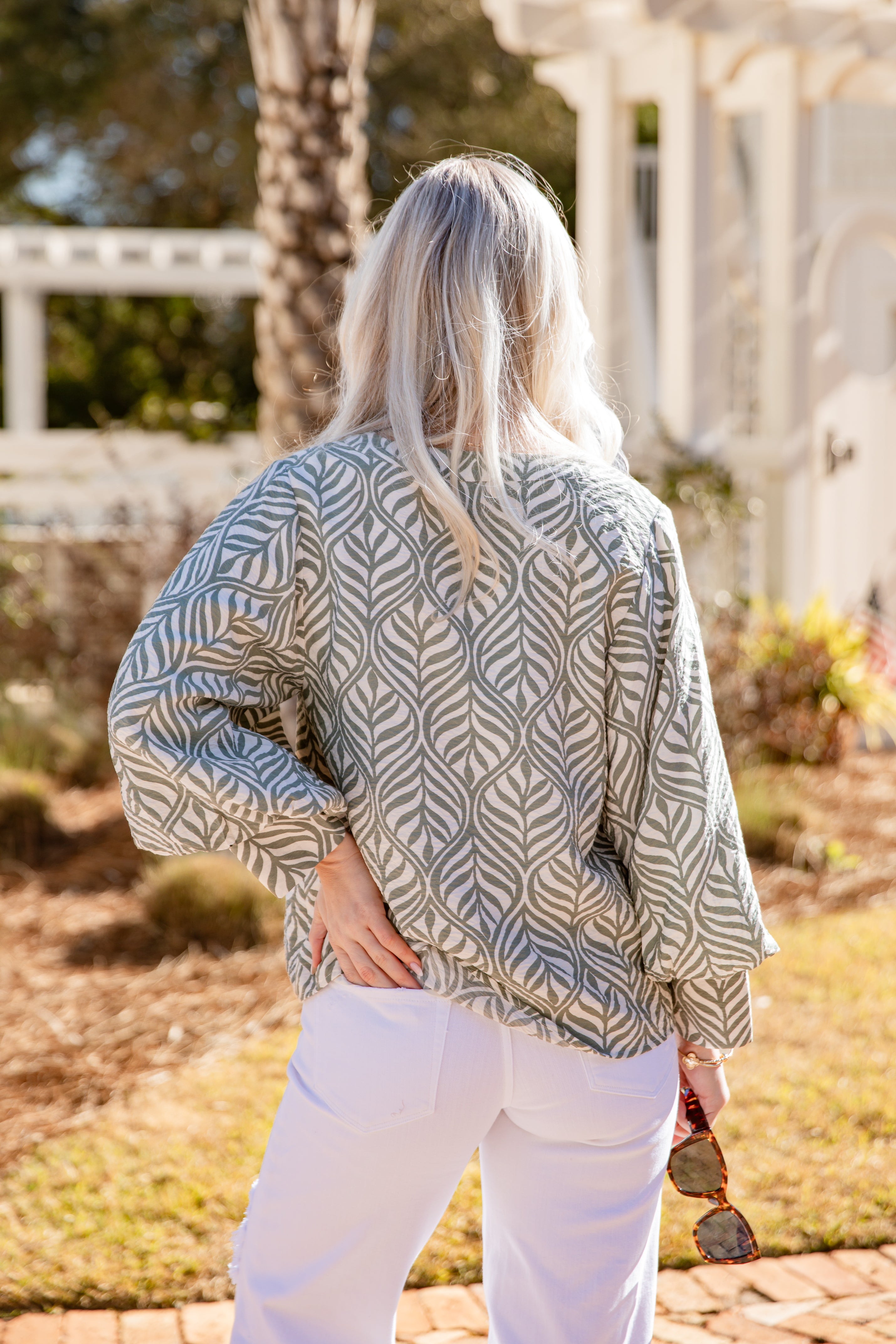 Woman wearing a patterned top and white pants standing outdoors with trees and a building in the background.