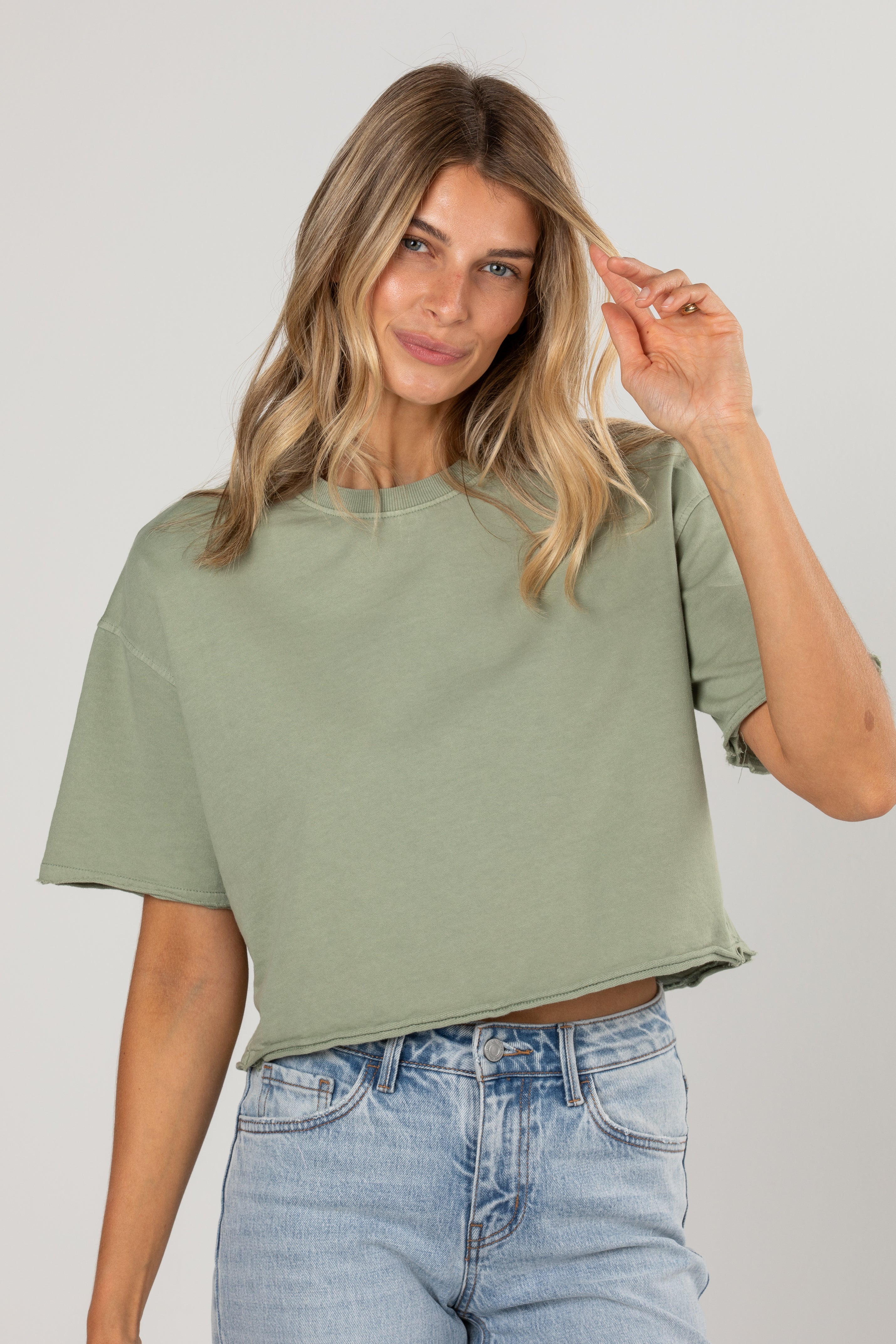 A woman wearing a cropped sage green t-shirt and light wash blue jeans, posing against a neutral light gray background.