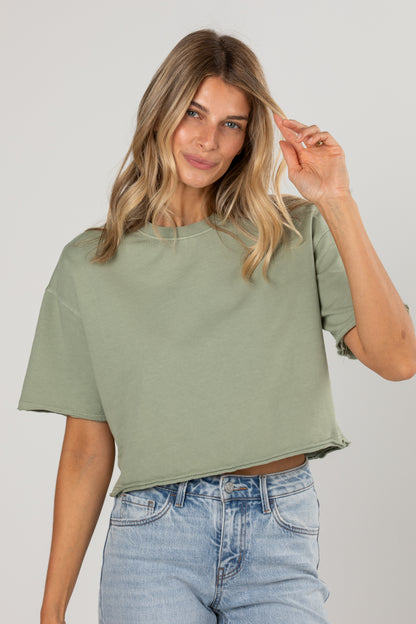 A woman wearing a cropped sage green t-shirt and light wash blue jeans, posing against a neutral light gray background.