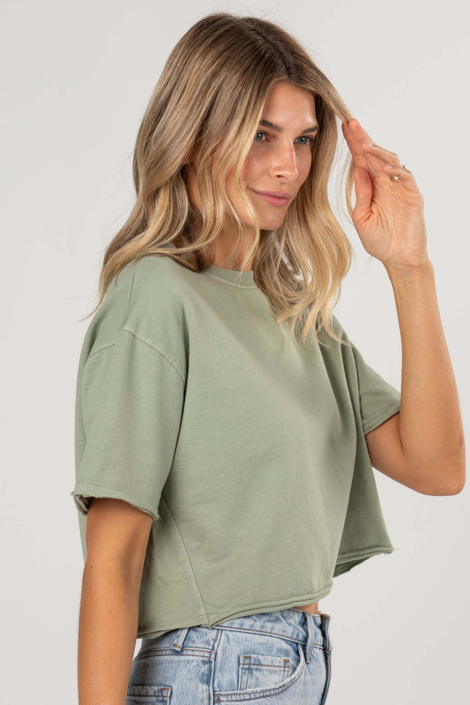 A woman wearing a sage green cropped short-sleeve t-shirt and light wash blue jeans, photographed against a solid light gray background.
