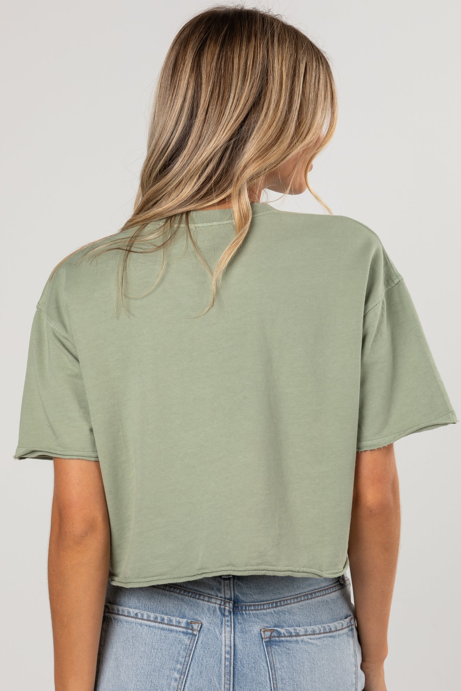 A woman viewed from the back wearing an oversized sage green short-sleeve crop top and light blue jeans against a plain light gray studio background.