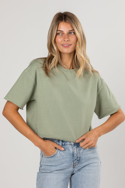 A woman wearing a light sage green short-sleeve crop top and light blue jeans, standing against a neutral light gray studio background.