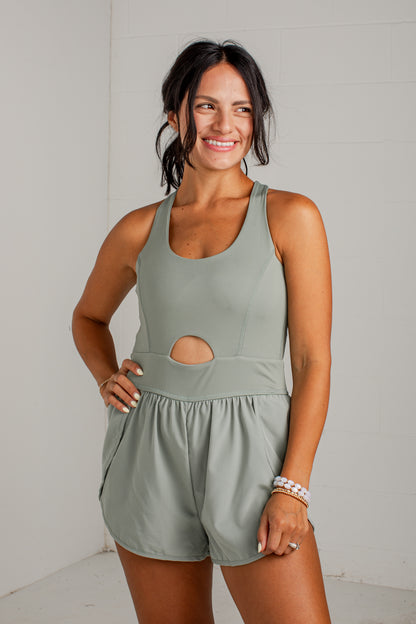 A woman modeling a sage green sleeveless romper with a cutout at the waist, standing against a white tiled wall.