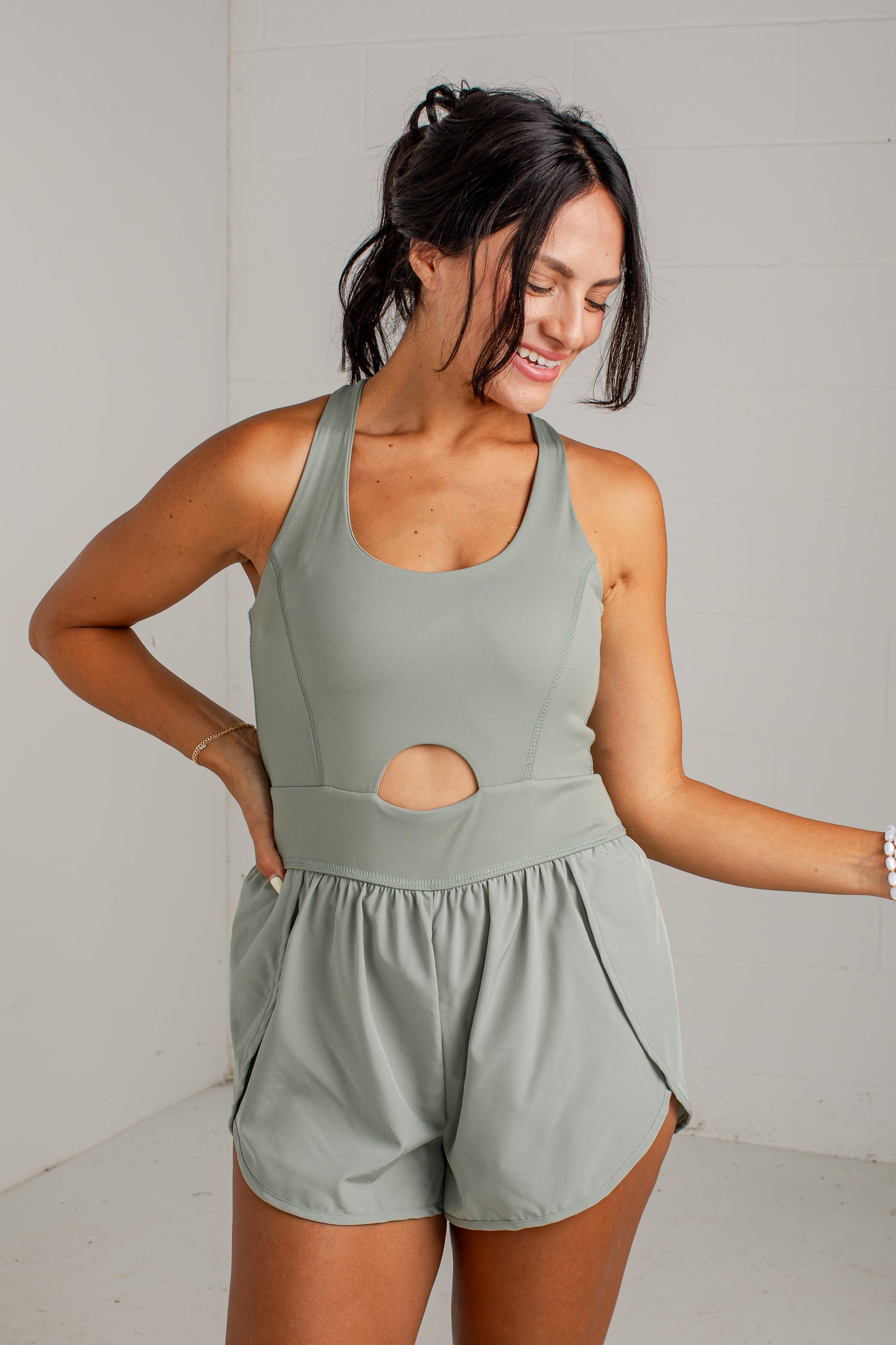 A woman modeling a sage green athletic romper with a scoop neckline, a central cutout, and matching shorts, set against a light gray brick wall.