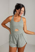 A woman modeling a sage green athletic romper with a scoop neckline, a central cutout, and matching shorts, set against a light gray brick wall.