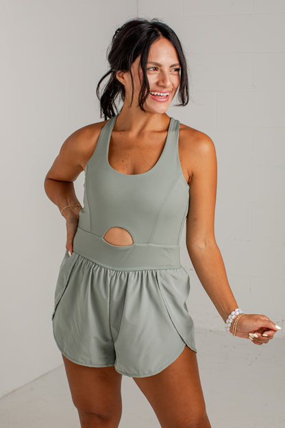 A woman modeling a sage green sleeveless romper with a side cutout, standing against a light gray brick wall.