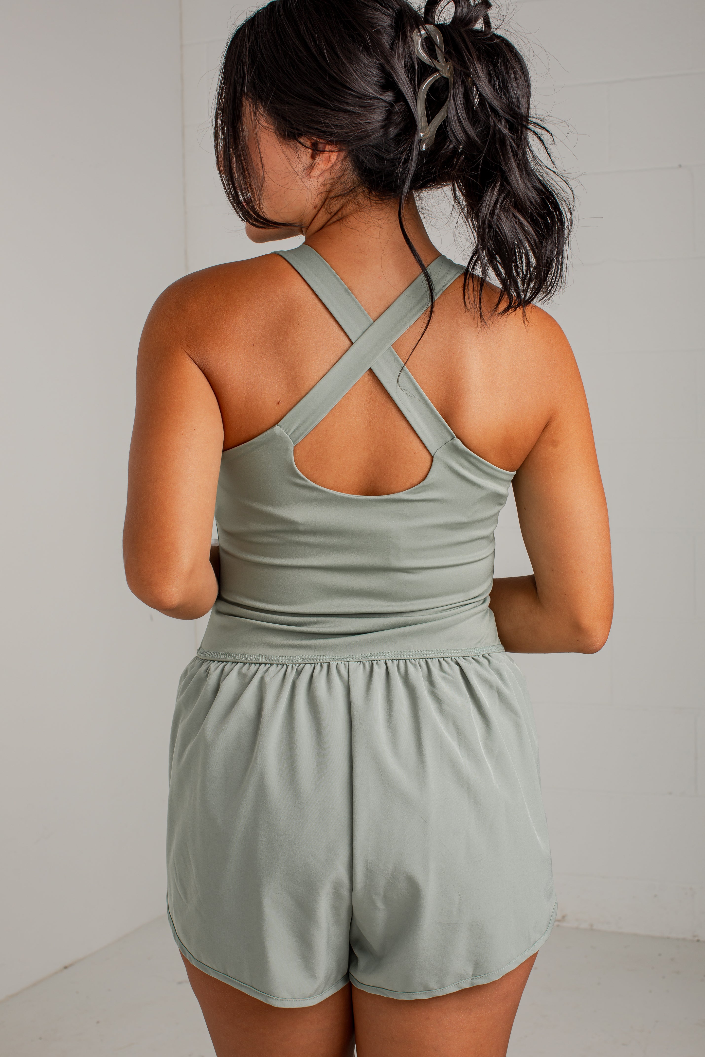 A woman shown from the back wearing a sage green athletic romper with crisscross shoulder straps and short legs, photographed against a plain light gray wall.
