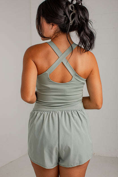 A woman shown from the back wearing a sage green athletic romper with crisscross shoulder straps and short legs, photographed against a plain light gray wall.