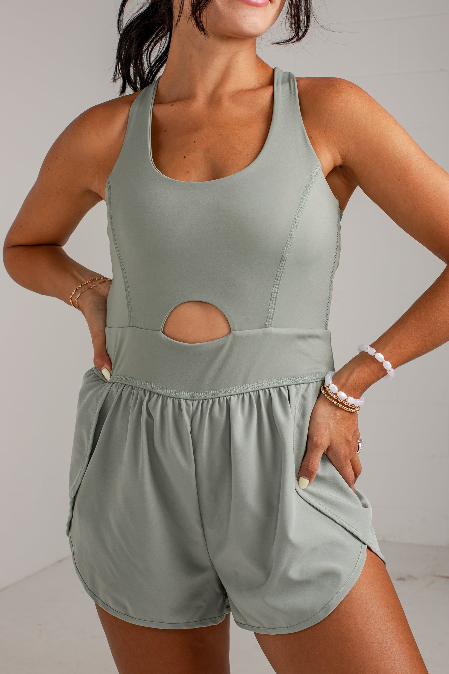 A woman wearing a sage green sleeveless romper with a scoop neckline, a side cutout, and matching shorts with side pockets, posed against a light gray wall.