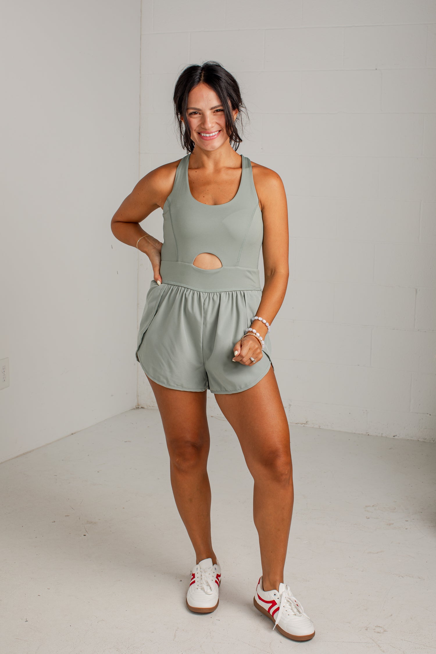 A woman modeling a sage green athletic romper with a cutout at the waist, paired with white sneakers, standing against a white brick wall.