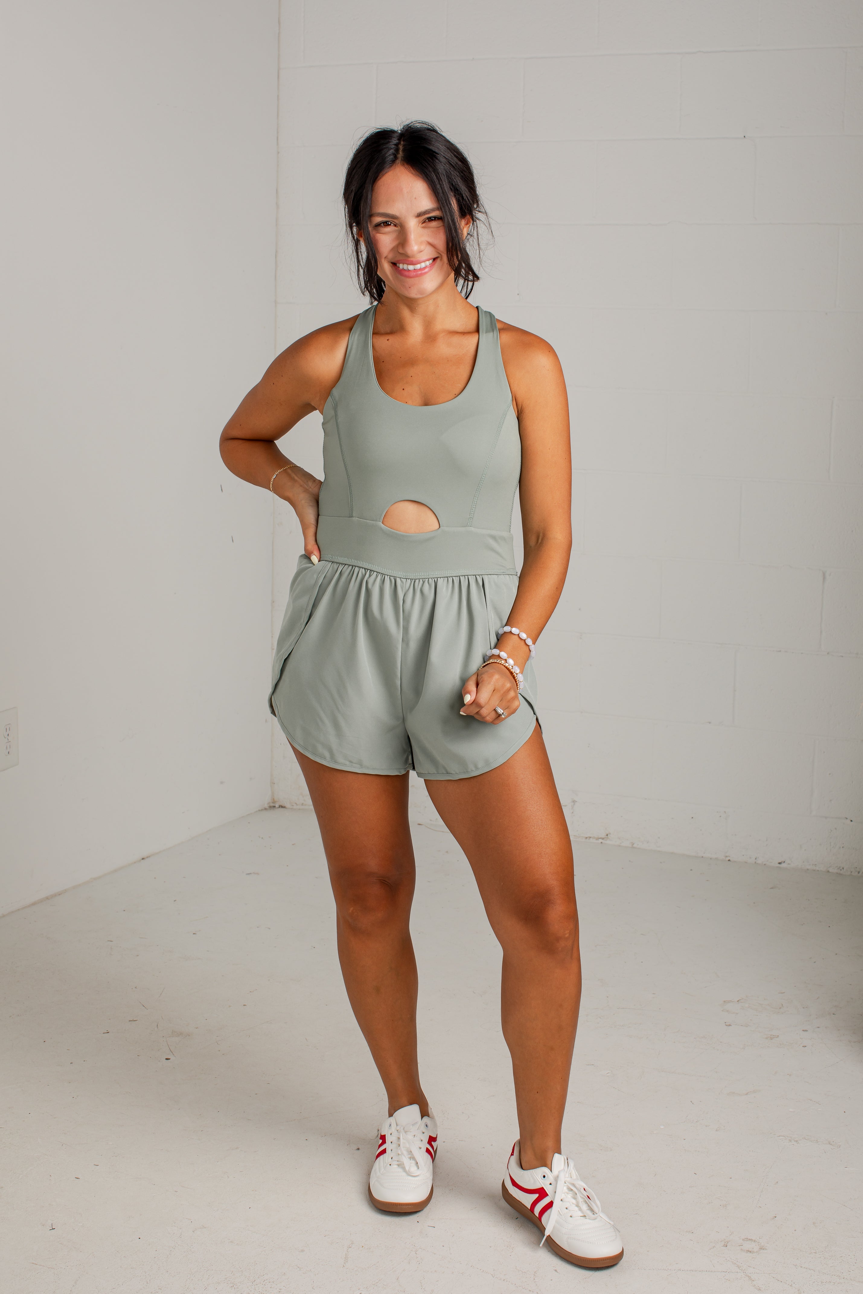 A woman modeling a sage green athletic romper with a cutout at the waist, paired with white sneakers, standing against a white brick wall.
