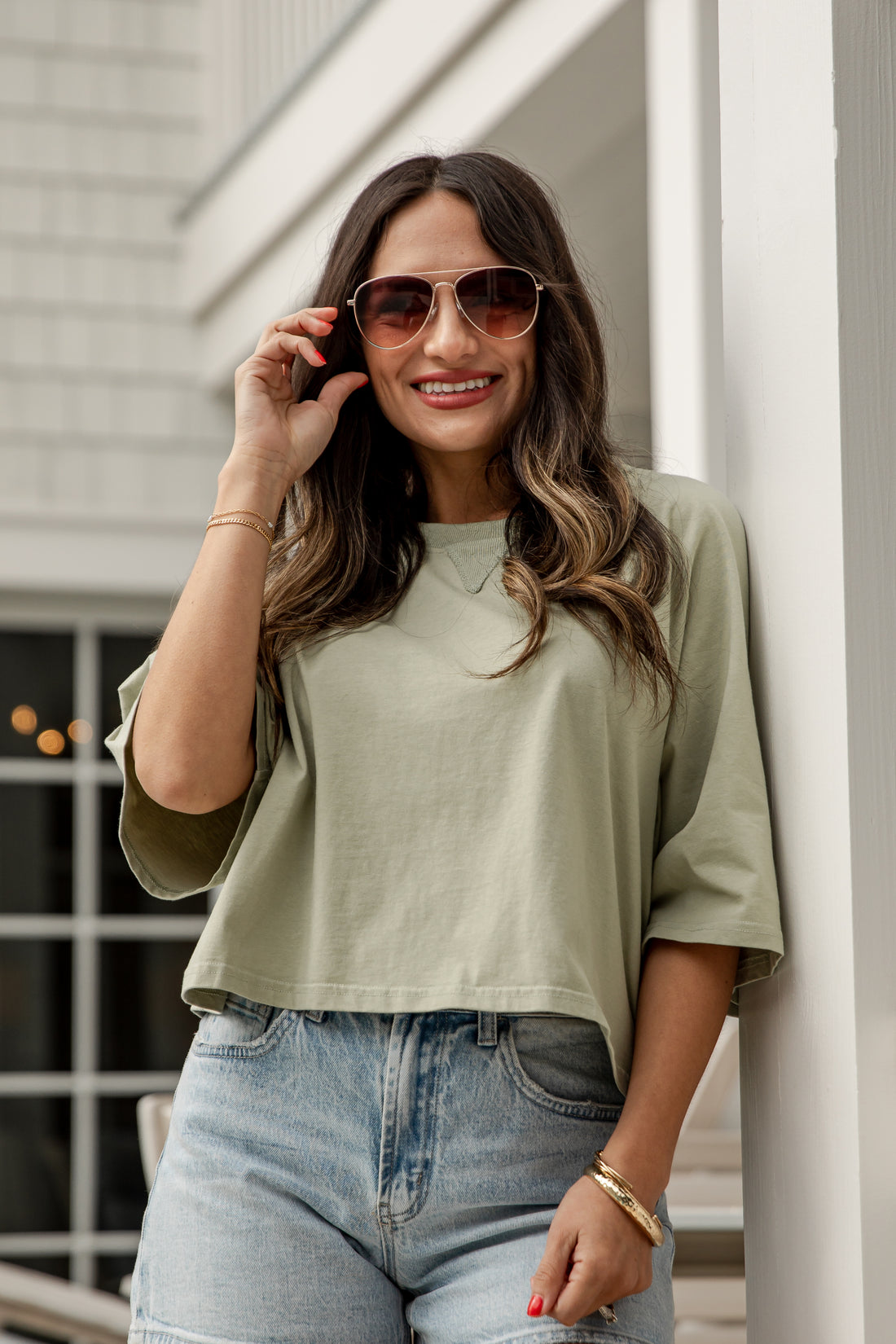 Woman wearing sunglasses and a green top, standing against a white wall.