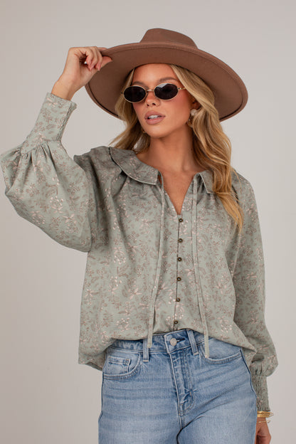 A woman wearing a light sage green long-sleeve button-up blouse with a subtle floral pattern, styled with a brown wide-brim hat, sunglasses, and light-wash blue jeans against a solid light gray background.