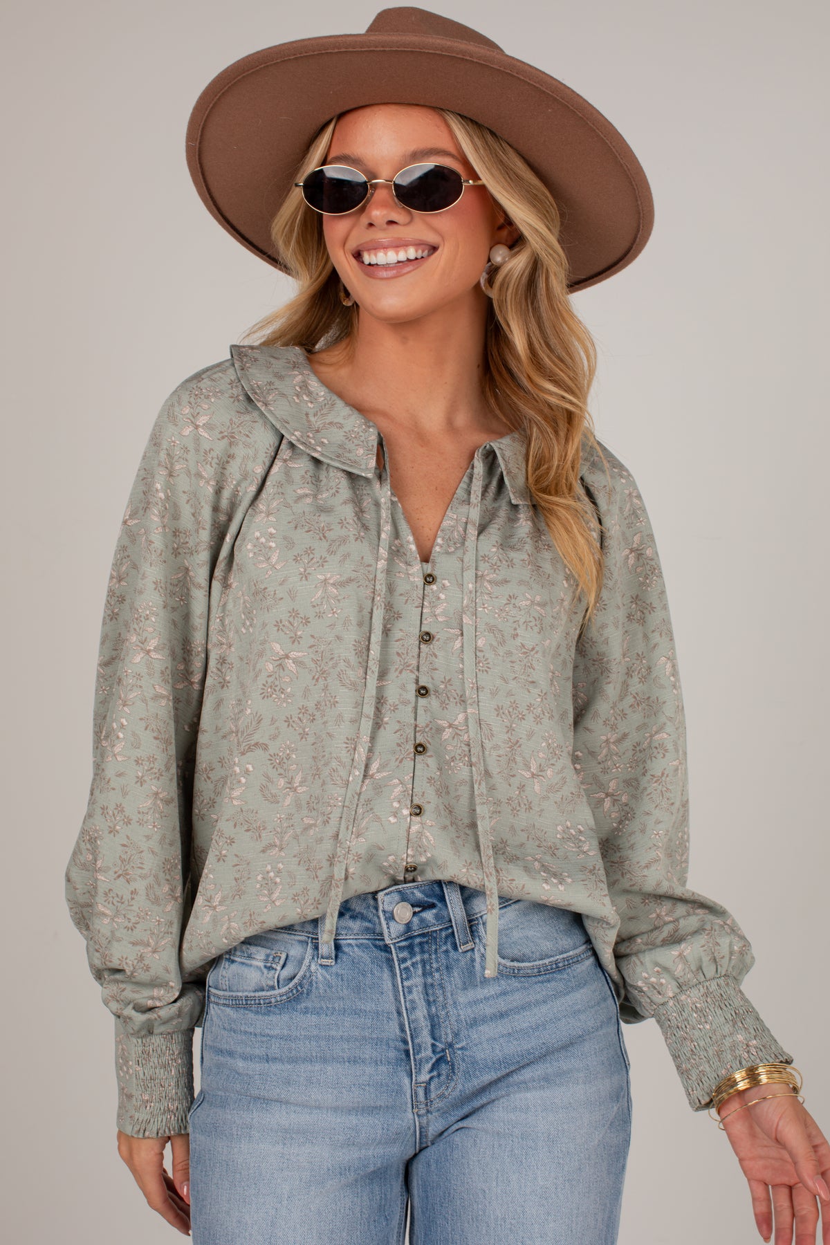 A woman wearing a light sage green long-sleeve button-up blouse with a subtle floral pattern, styled with blue jeans, a brown wide-brim hat, and sunglasses against a neutral studio background.
