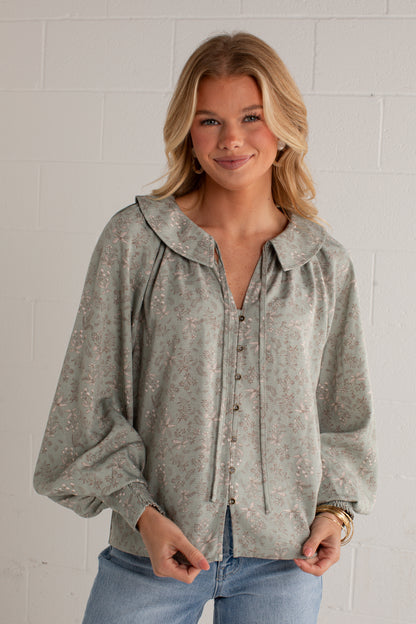 A woman wearing a sage green long-sleeve blouse with a delicate floral pattern, paired with blue jeans, standing against a white brick wall.