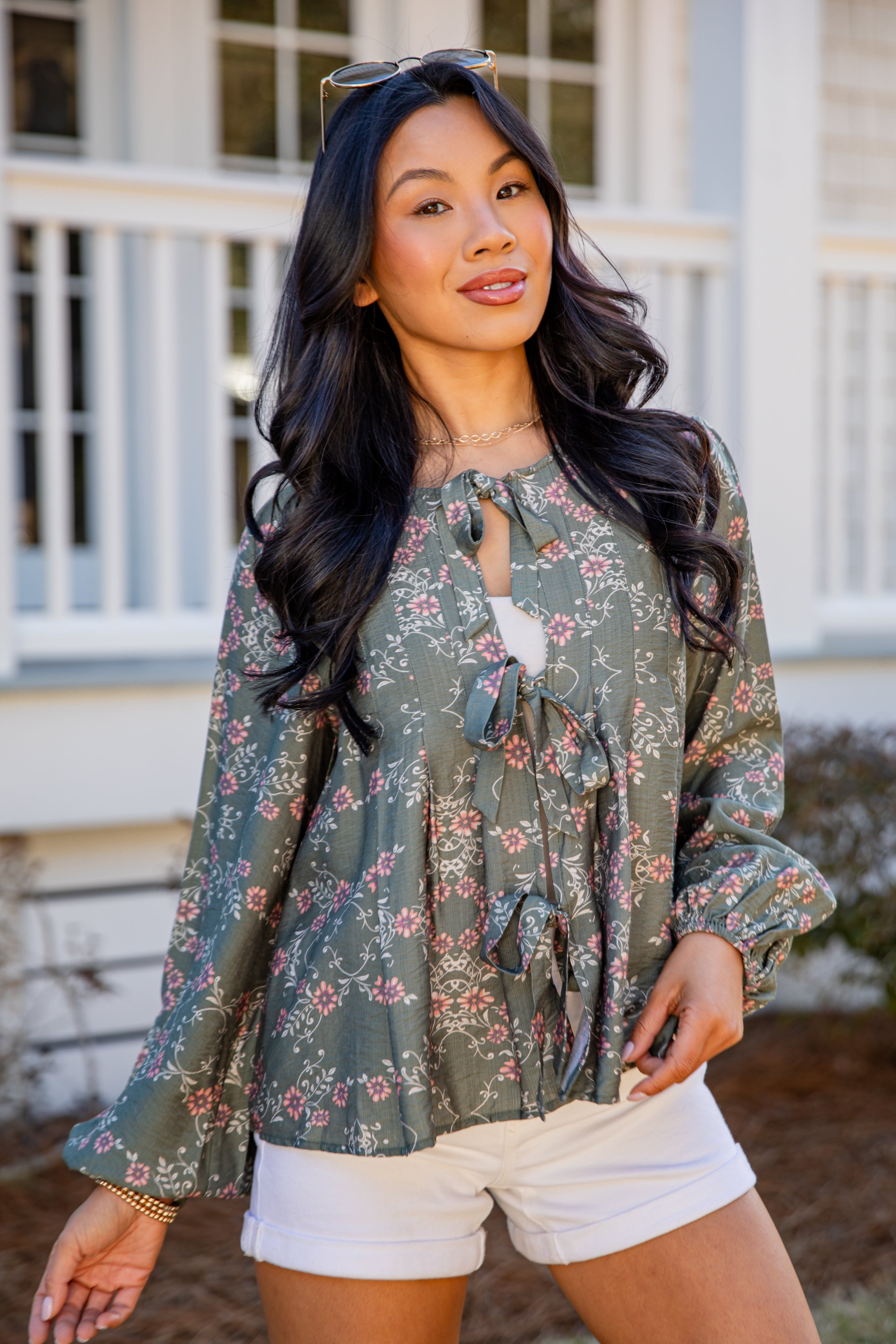 Woman wearing a floral blouse and white shorts standing in front of a house.