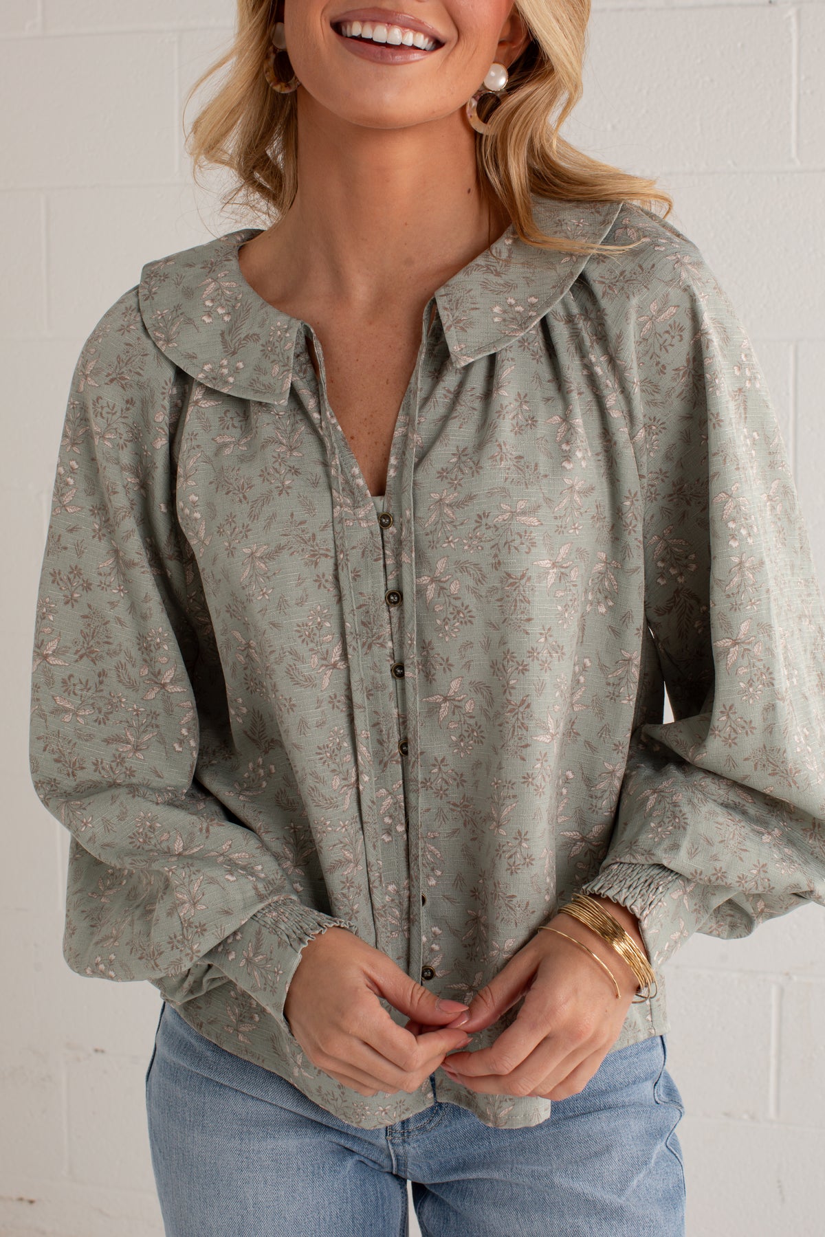 A woman wearing a sage green long-sleeve blouse with a delicate pink floral pattern, a large collar, and a button-down front, paired with light blue jeans.