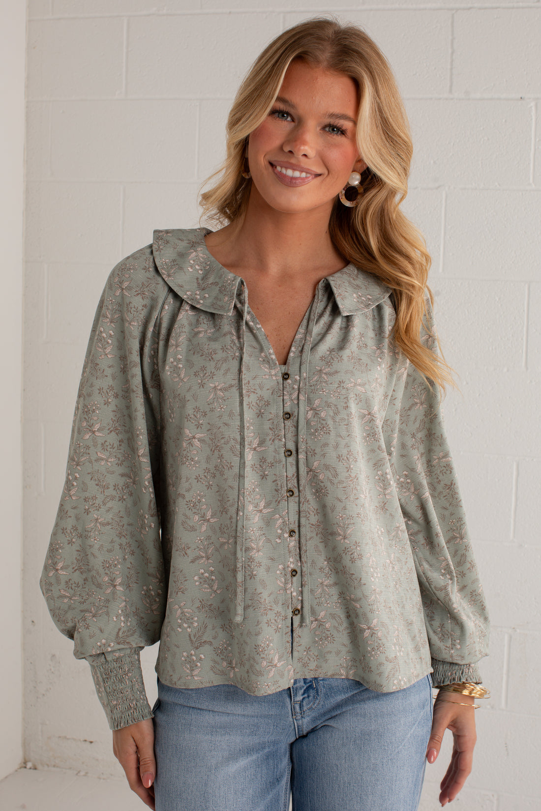 A woman wearing a sage green long-sleeve blouse with a delicate pink floral pattern, a wide collar, and a button-down front, paired with blue jeans against a white brick wall.