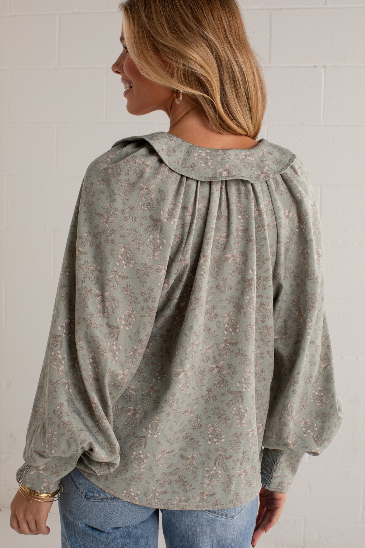 A woman shown from the back wearing a sage green long-sleeve blouse with a delicate floral pattern and a gathered collar, paired with blue jeans against a white brick wall.