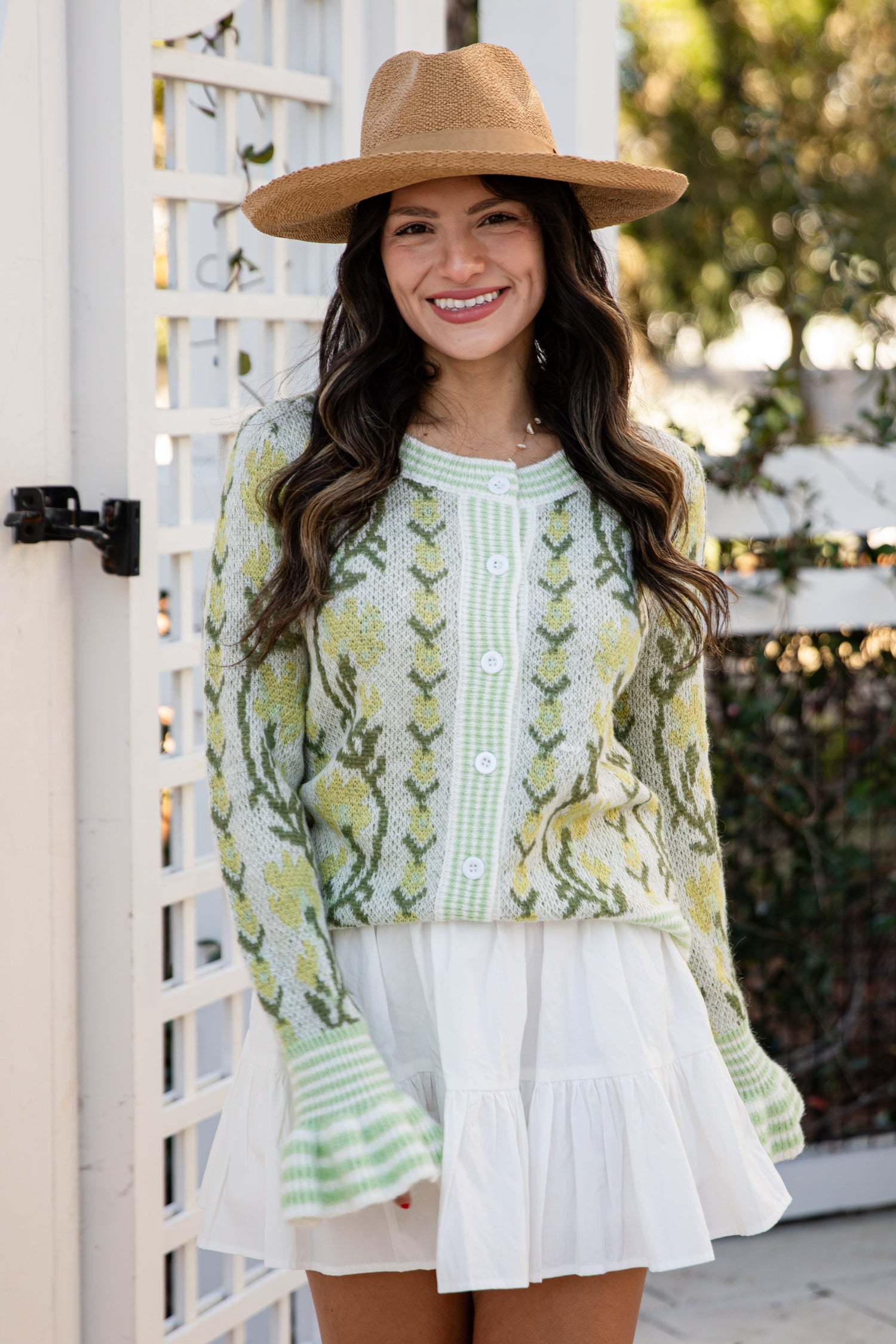 Woman wearing a green patterned top and white skirt with a brown hat, standing outdoors.