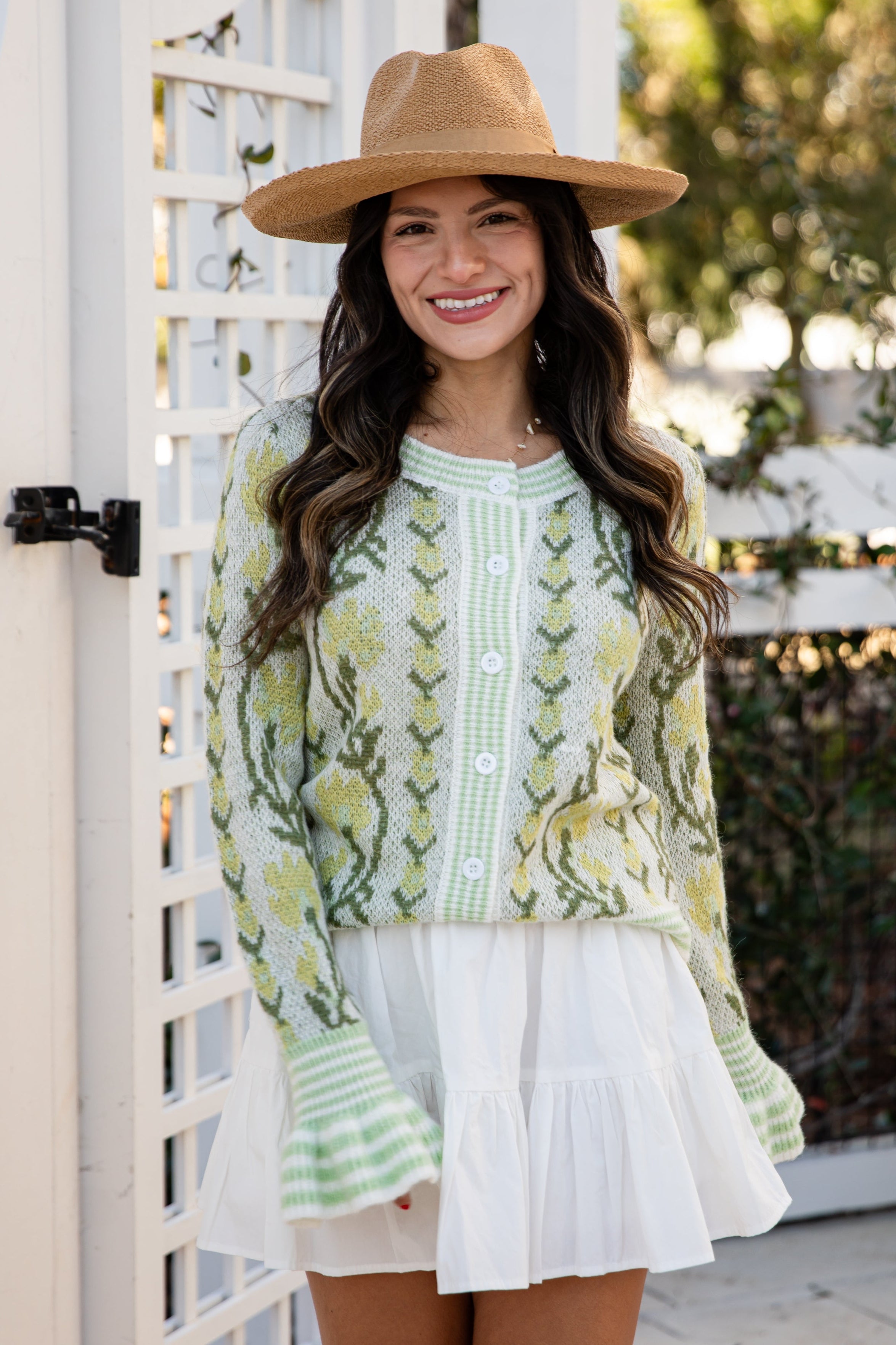 Woman wearing a green patterned top and white skirt with a brown hat, standing outdoors.