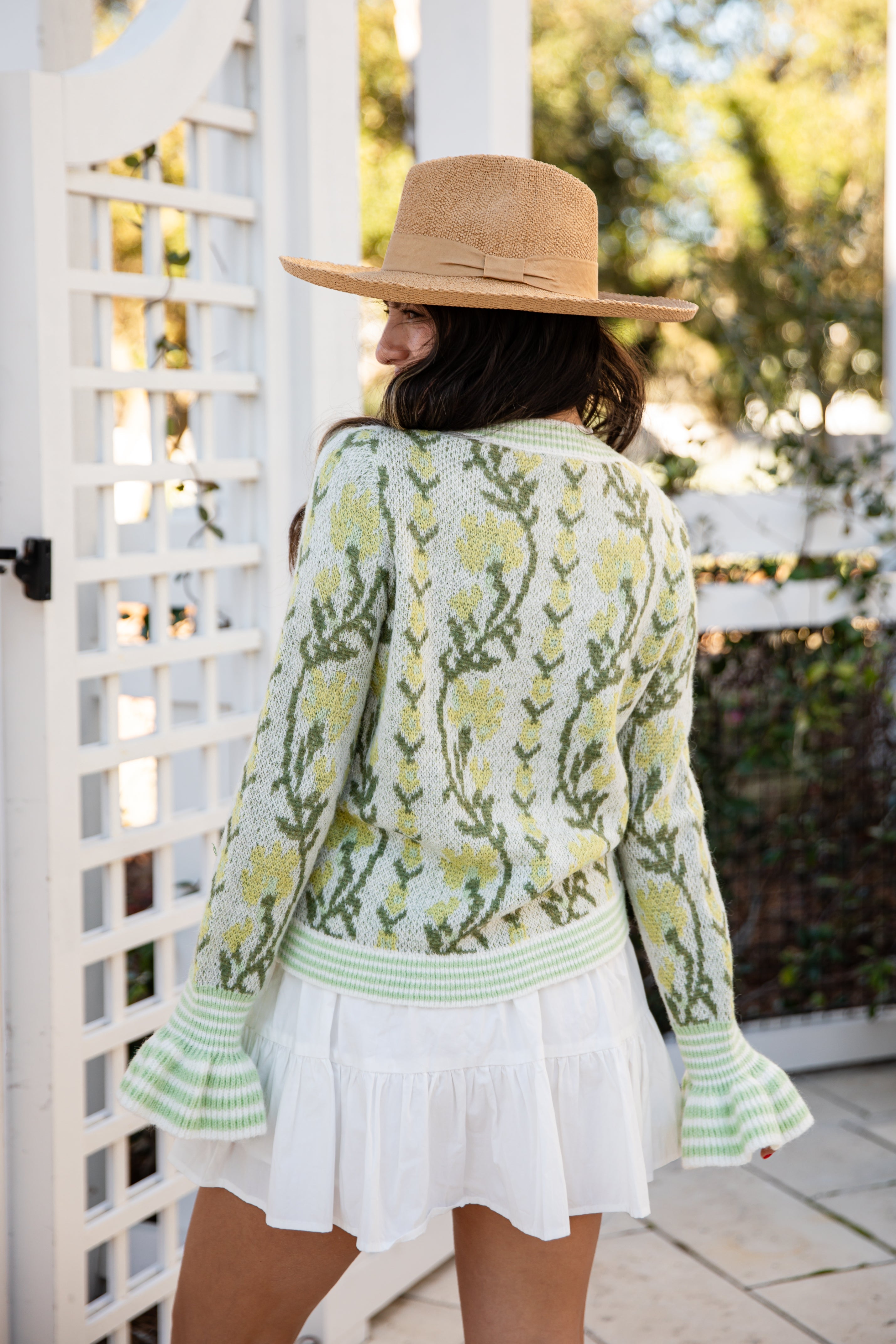 Woman wearing a floral cardigan and white dress with a straw hat, standing in a garden.