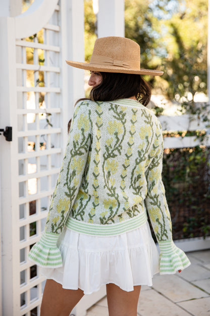 Woman wearing a floral cardigan and white dress with a straw hat, standing in a garden.