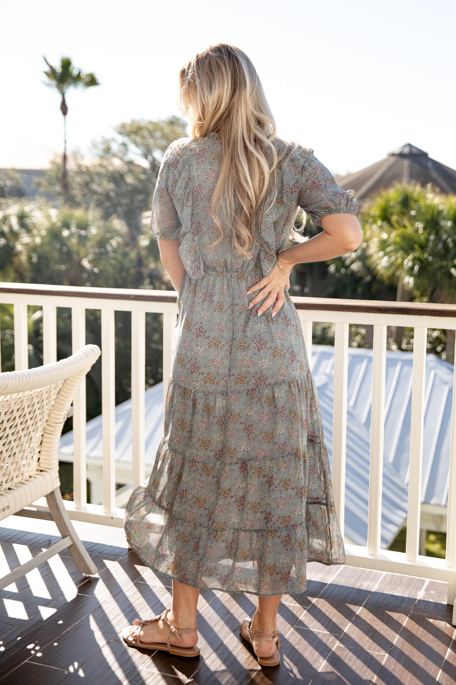 Woman in a floral dress standing on a balcony with trees and a building in the background