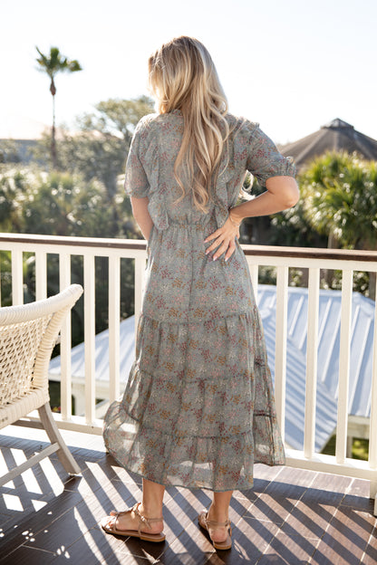 Woman in a floral dress standing on a balcony with trees and a building in the background
