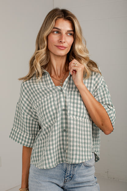 A woman wearing a sage green and white gingham button-down shirt with rolled sleeves, paired with light-wash blue jeans, standing against a light gray wall.