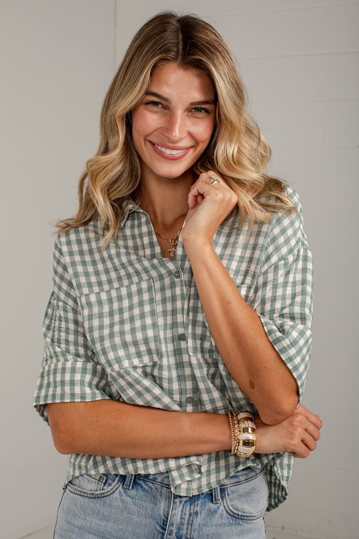 A smiling woman with blonde hair wearing a sage green and white gingham button-down shirt and light-wash blue jeans, posed against a light gray wall.