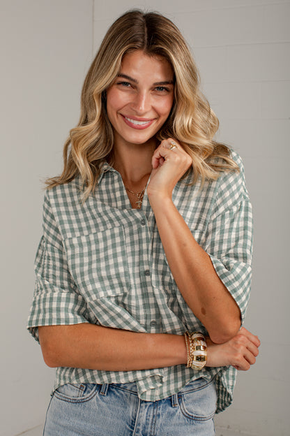 A smiling woman with blonde hair wearing a sage green and white gingham button-down shirt and light-wash blue jeans, posed against a light gray wall.