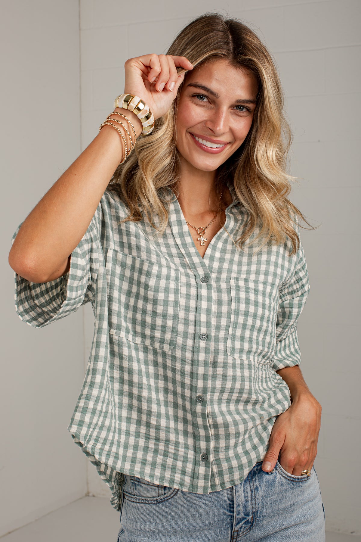 A woman wearing a green and white gingham button-down shirt with rolled-up sleeves and light-wash blue jeans, posing against a light gray wall.
