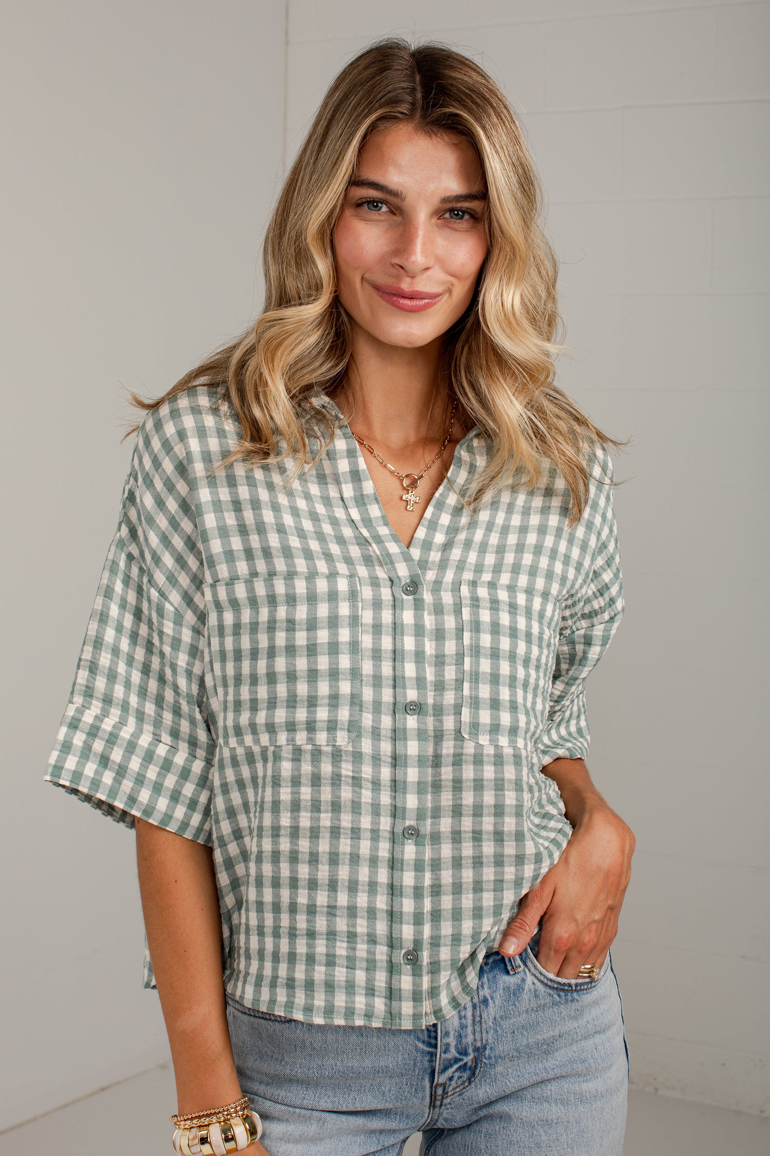 A woman wearing a green and white gingham short-sleeve button-up shirt with rolled cuffs, paired with light-wash blue jeans, standing against a light gray wall.