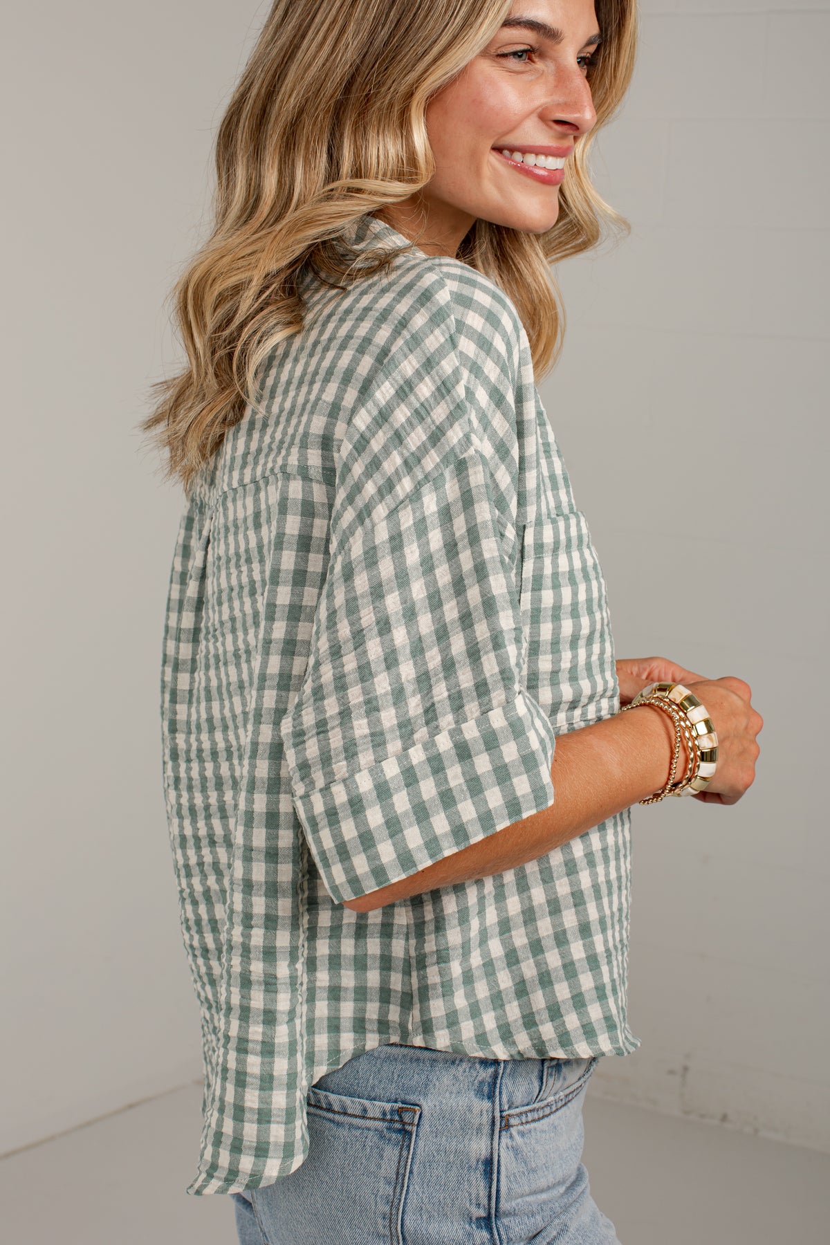 A woman wearing a sage green and white gingham check shirt with rolled-up sleeves, paired with light-wash blue jeans, shown from a side-back angle against a neutral studio background.