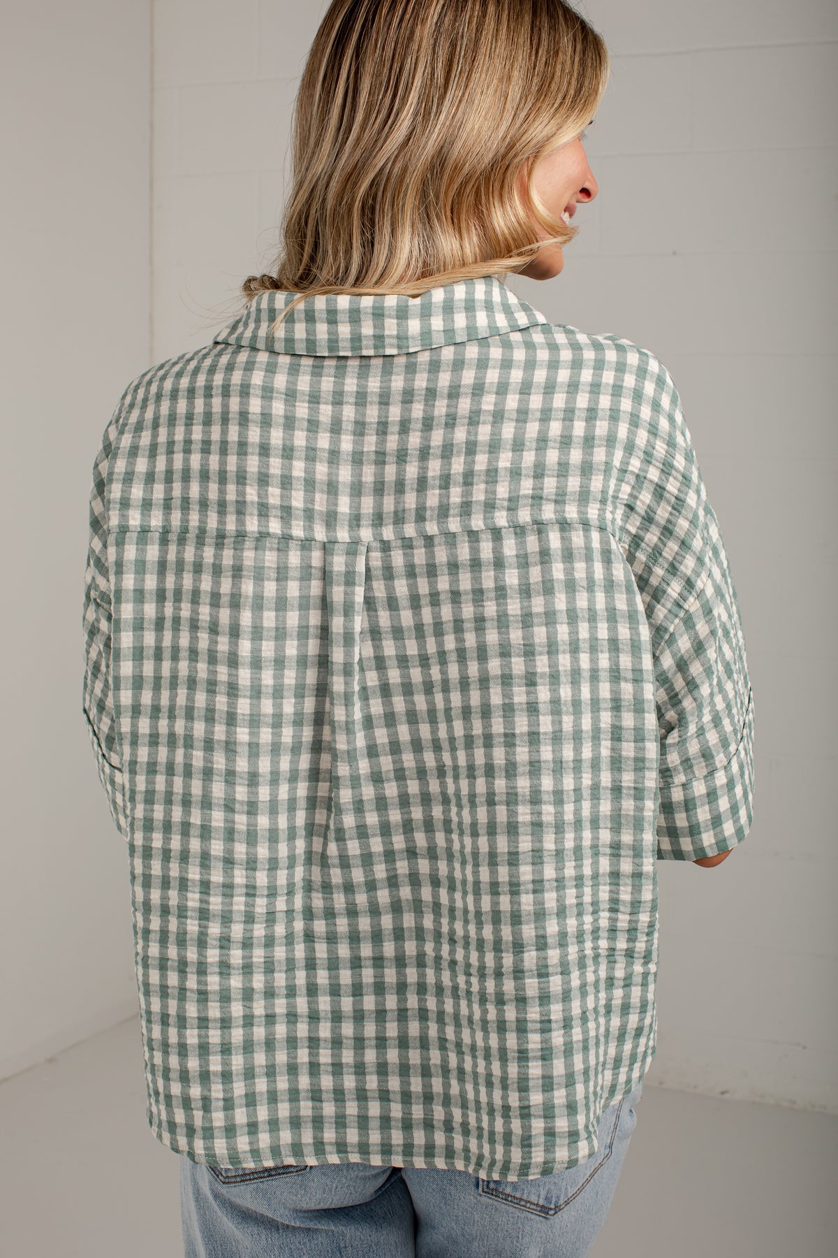 A woman shown from the back wearing a green and white gingham check shirt with light blue jeans, set against a plain light gray wall.