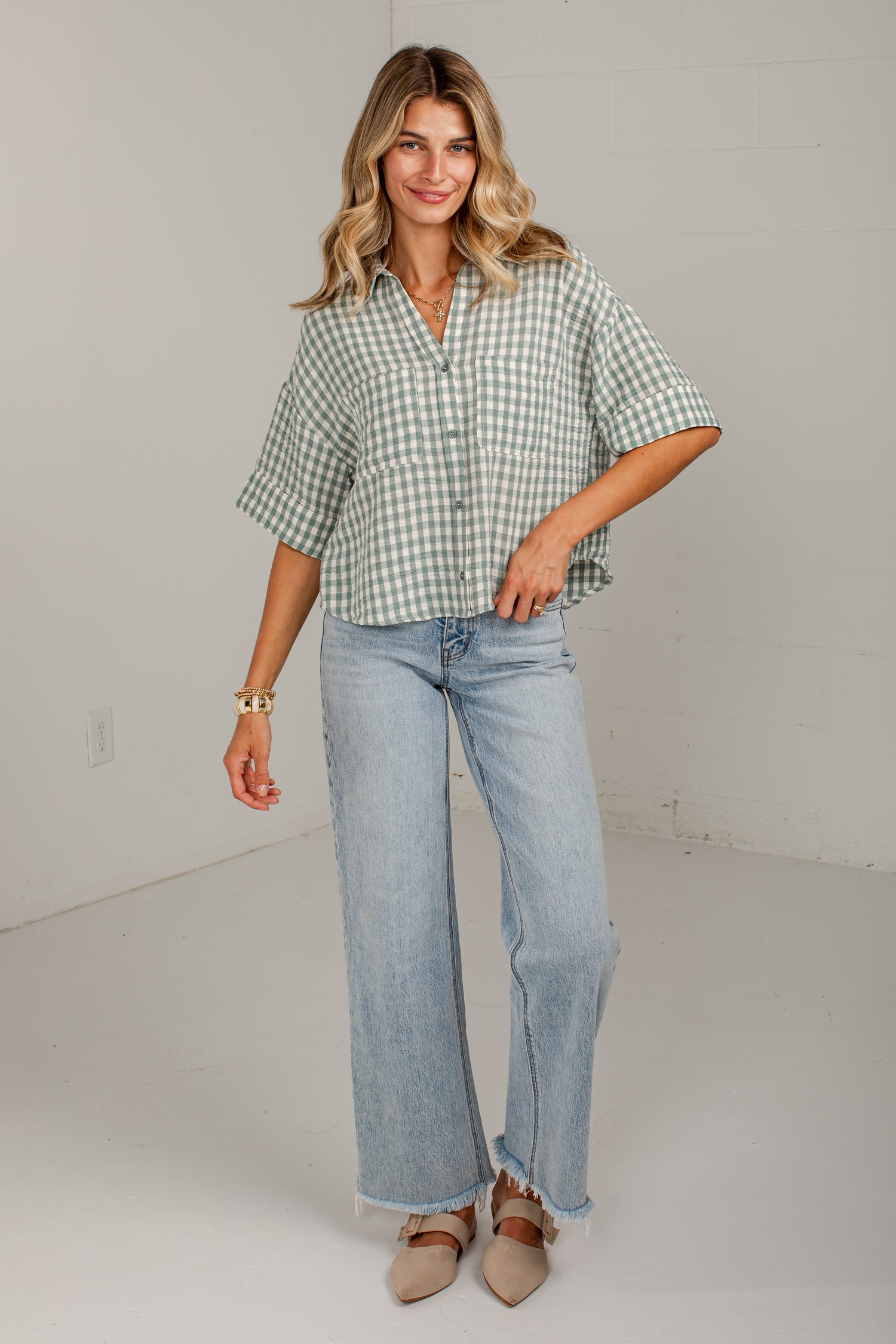 A woman modeling a light green and white gingham short-sleeve button-up shirt paired with light-wash wide-leg jeans and beige pointed-toe flats, standing against a plain off-white studio wall.