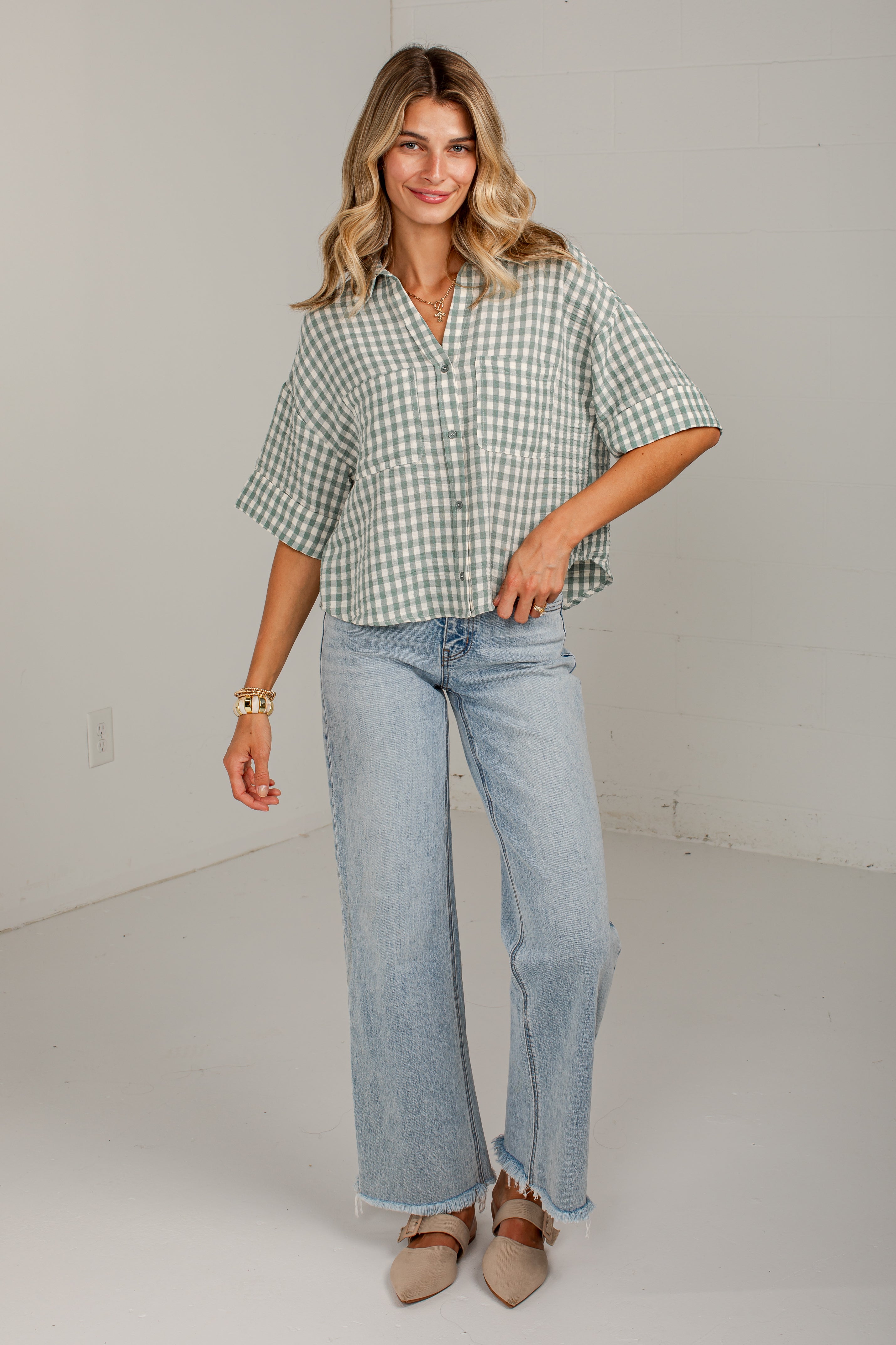 A woman modeling a light green and white gingham short-sleeve button-up shirt paired with light-wash wide-leg jeans and beige pointed-toe flats, standing against a plain off-white studio wall.