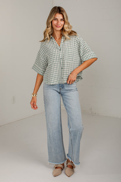 A woman modeling a light green and white gingham short-sleeve button-up shirt paired with light-wash wide-leg jeans and beige pointed-toe flats, standing against a plain off-white studio wall.