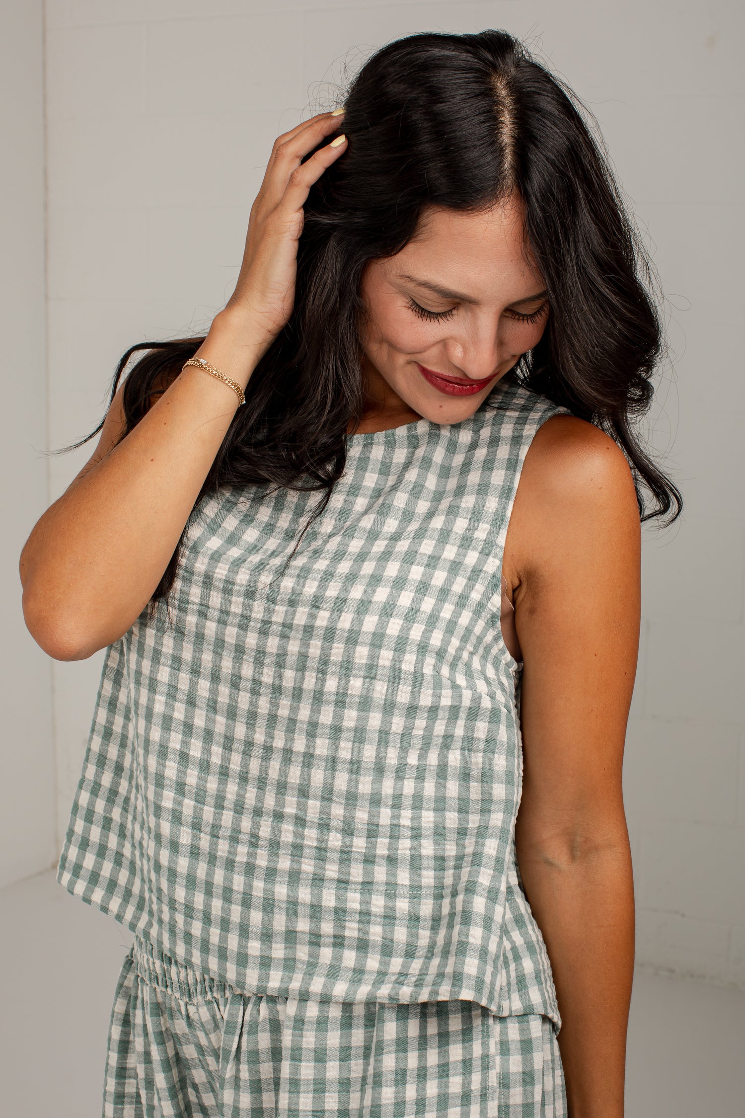 A woman wearing a sleeveless green and white gingham check top, shown from the chest down against a plain light gray wall.