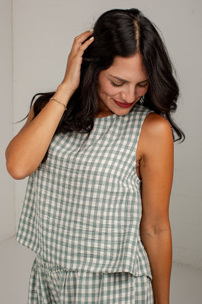 A woman wearing a sleeveless green and white gingham check top, shown from the chest down against a plain light gray wall.