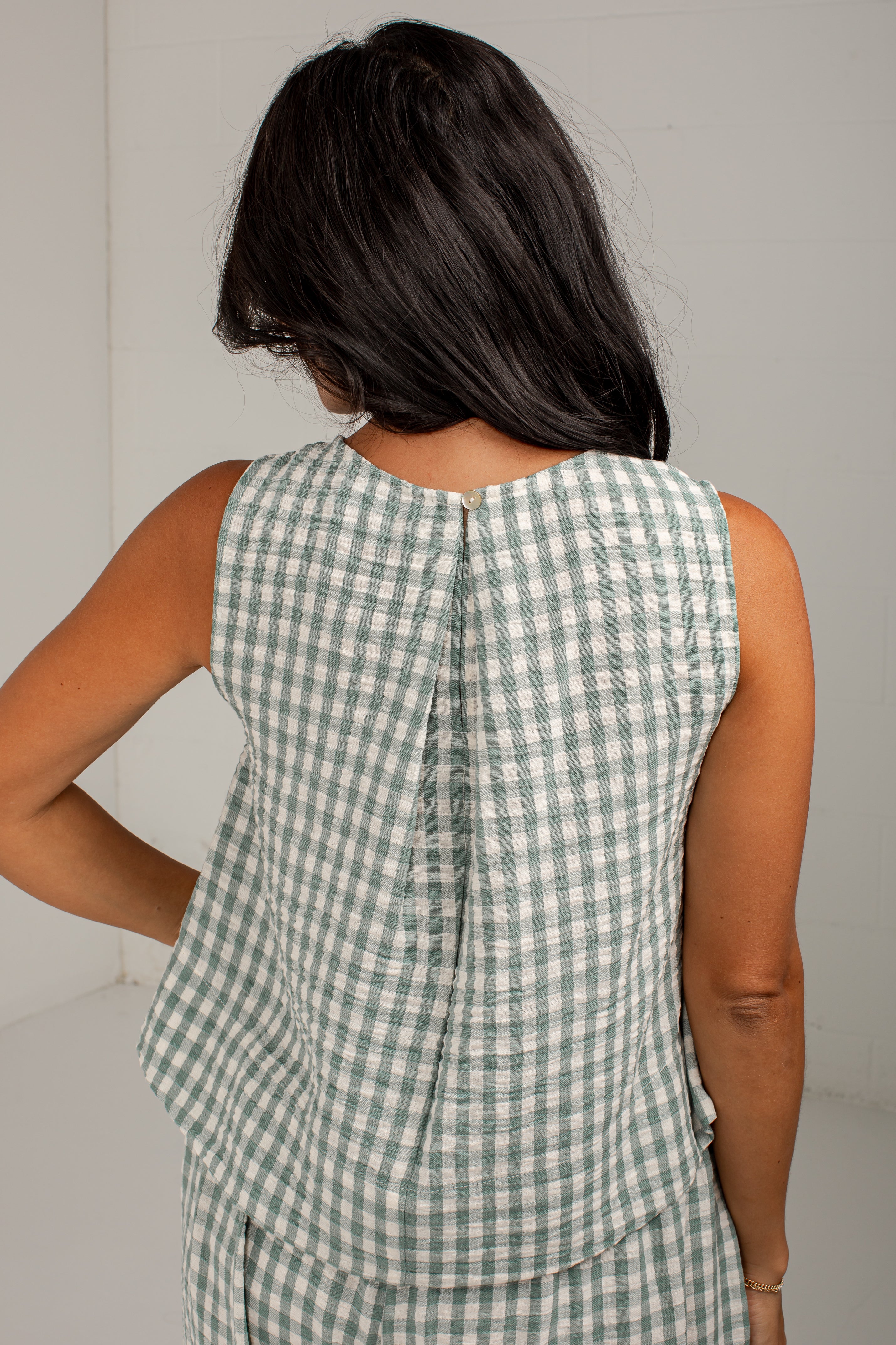A woman shown from behind wearing a sleeveless green and white gingham top with a gathered peplum back, photographed against a plain light gray wall.