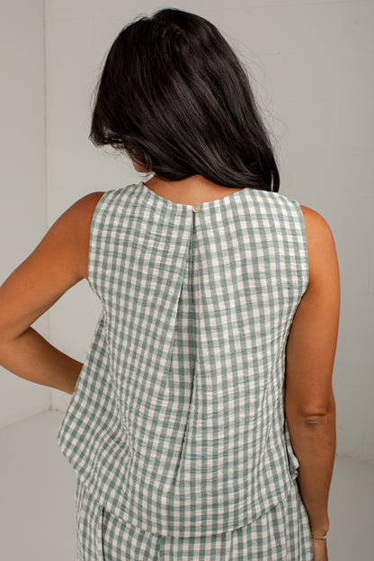 A woman shown from behind wearing a sleeveless green and white gingham top with a gathered peplum back, photographed against a plain light gray wall.