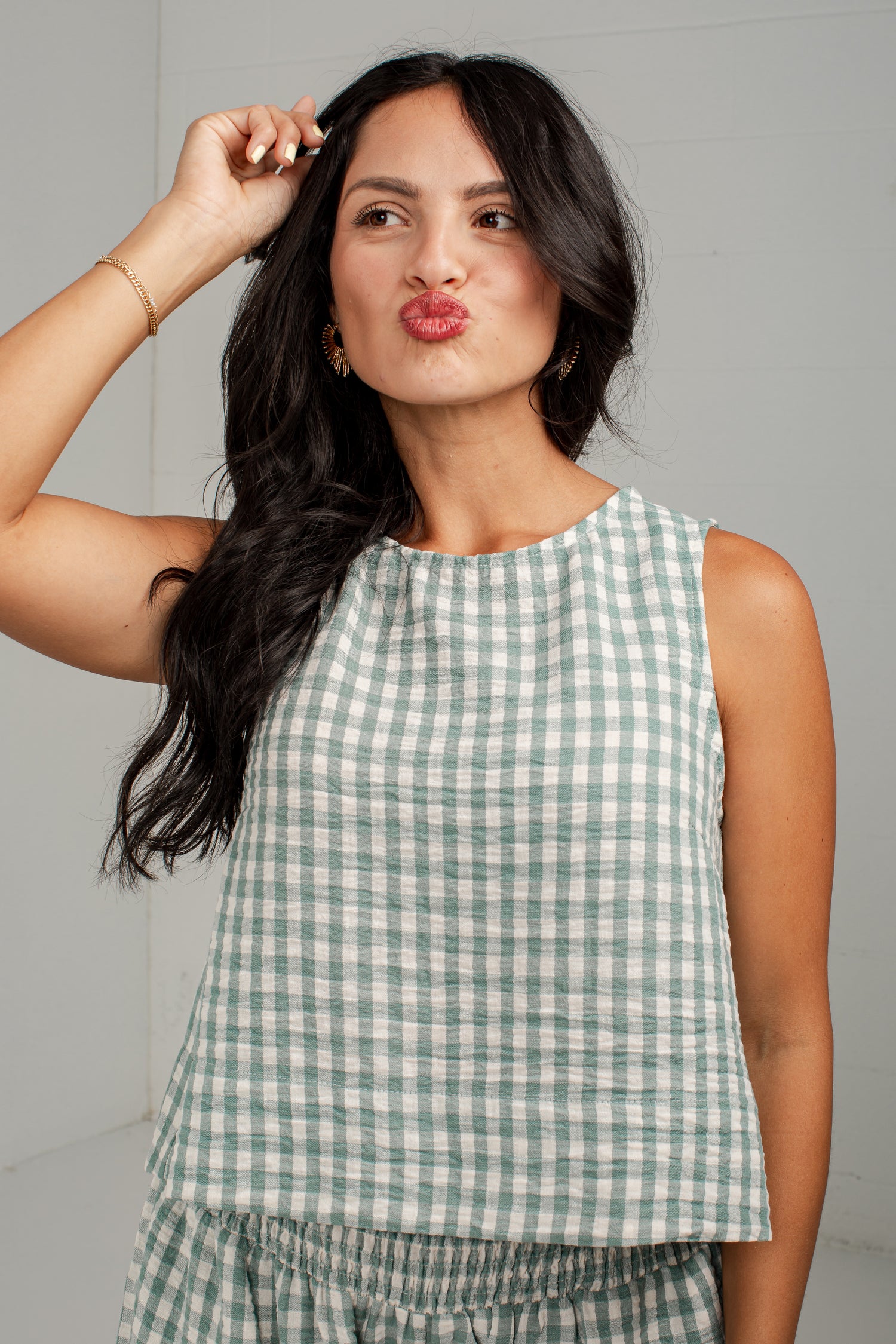 A woman wearing a sleeveless green and white gingham top, posing against a light gray studio background.