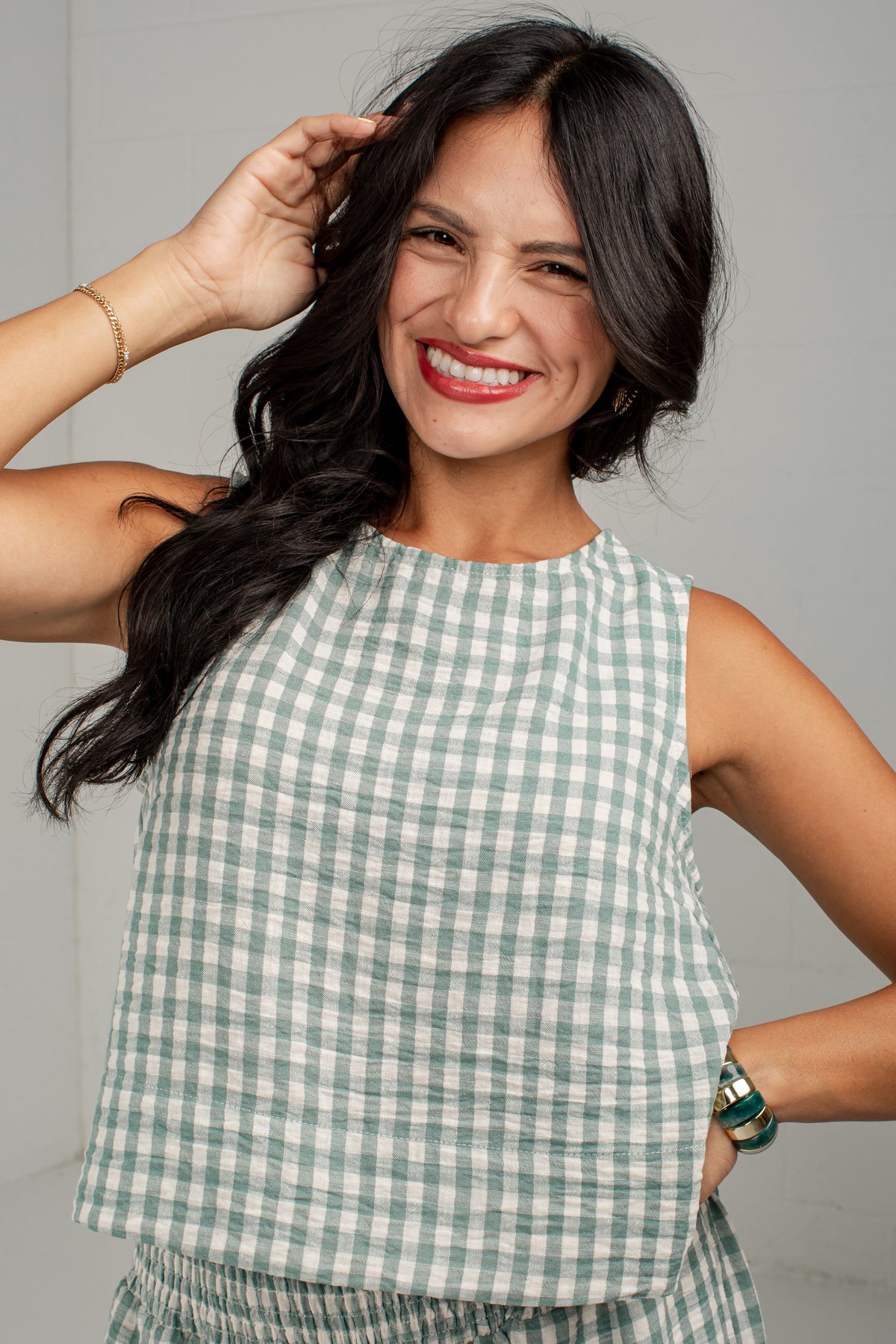 A smiling woman with long dark hair wearing a sleeveless light green and white gingham check top, posing against a neutral light gray background.