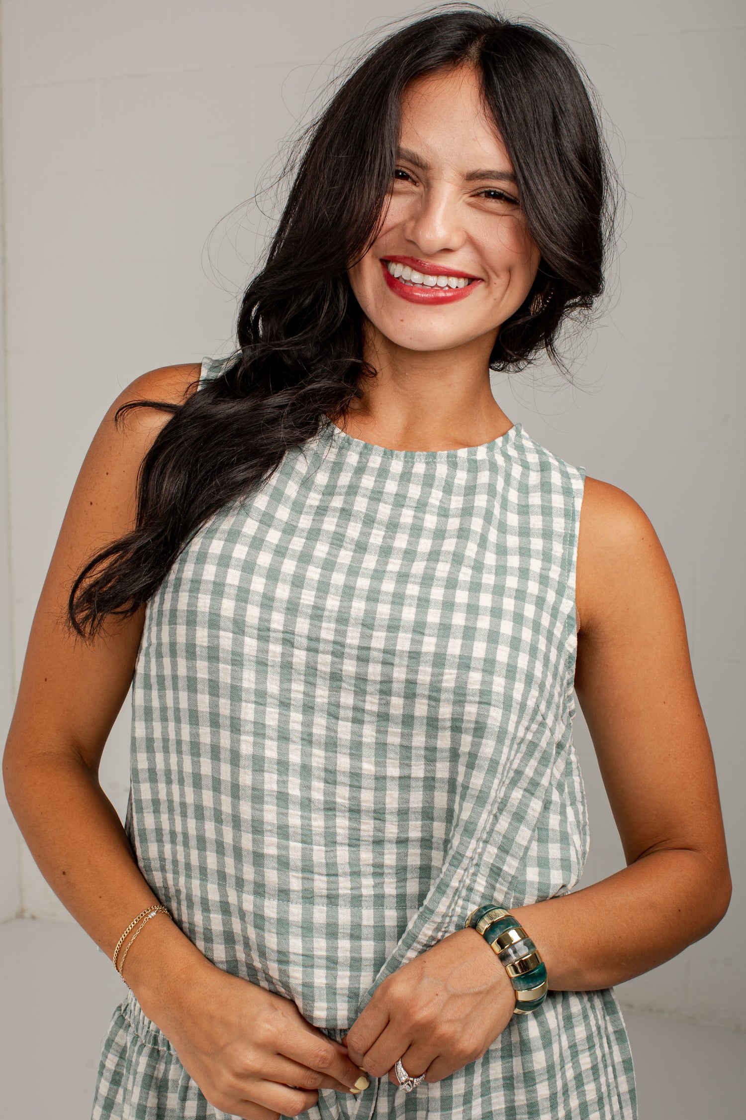 A smiling woman with long dark hair wearing a sleeveless green and white gingham dress, standing against a light gray background.