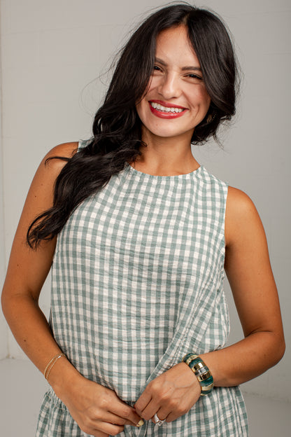 A smiling woman with long dark hair wearing a sleeveless green and white gingham dress, standing against a light gray background.