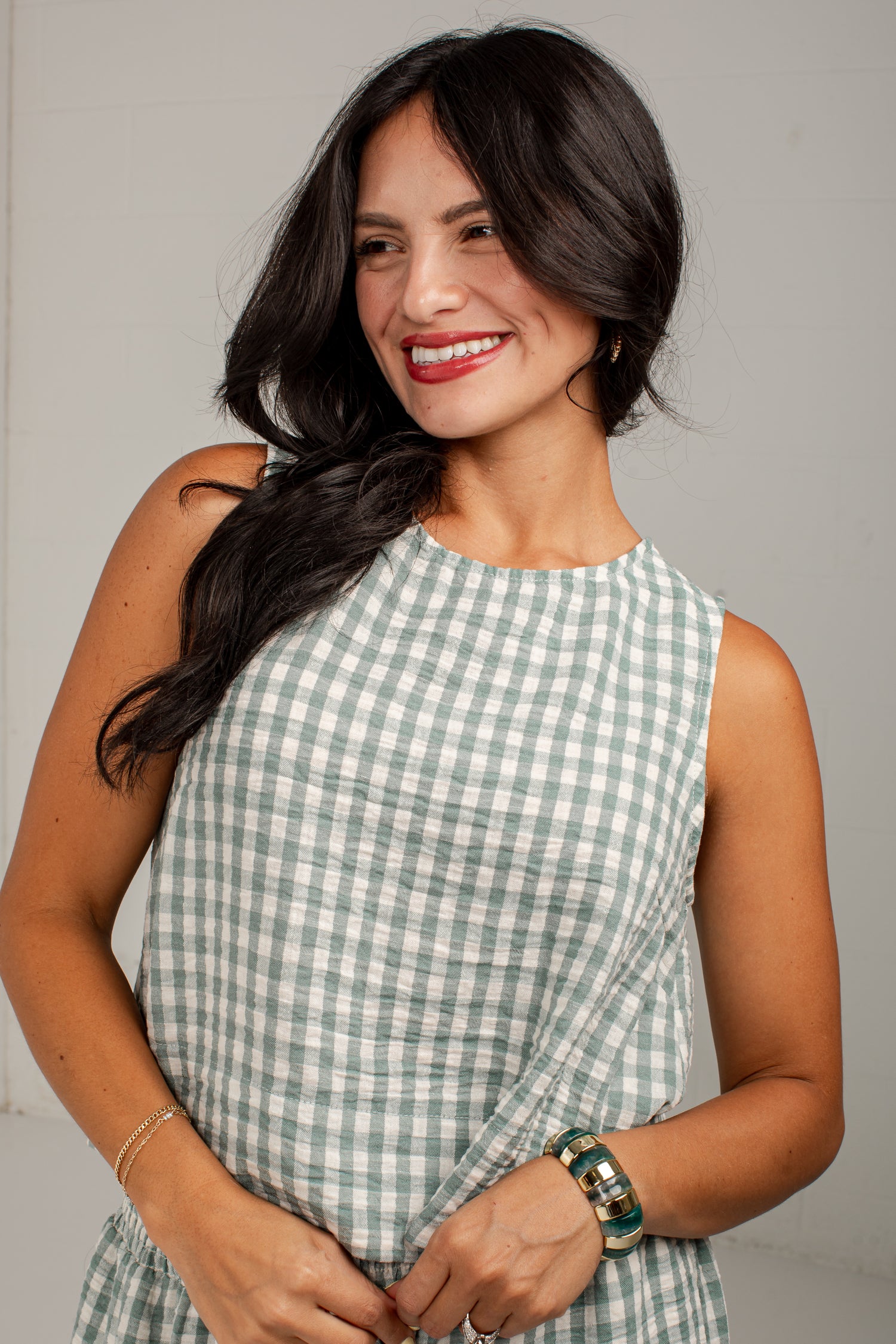 A woman with long dark hair smiling while wearing a sleeveless light green and white gingham dress against a plain light gray studio background.