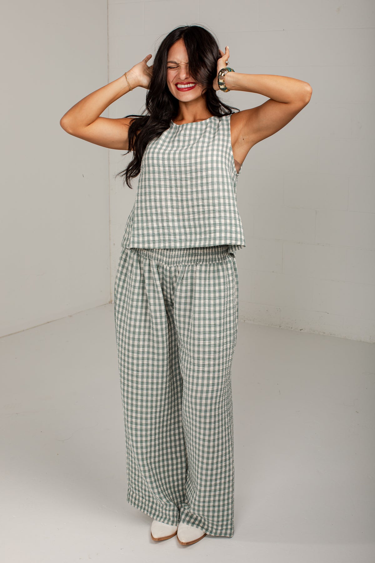A woman modeling a matching two-piece lounge set featuring a sleeveless crop top and wide-leg pants in a sage green and white gingham check pattern, set against a plain light gray studio background.